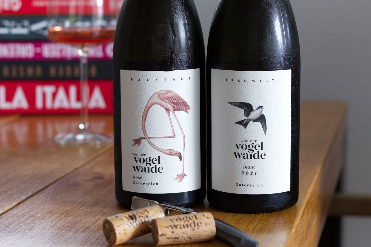 Zwei Flaschen Wein des Guts Von der Vogelwaide auf einem Holztisch. Die linke Flasche ist ein Rosé mit dem Namen Balztanz und zeigt einen Flamingo auf dem Etikett. Die rechte Flasche ist ein Weißwein mit dem Namen Frau Welt und zeigt einen fliegenden Vogel auf dem Etikett. Im Hintergrund sind ein Weinglas und Bücherstapel zu sehen. Vor den Flaschen liegen zwei Korken und ein Kellnermesser.