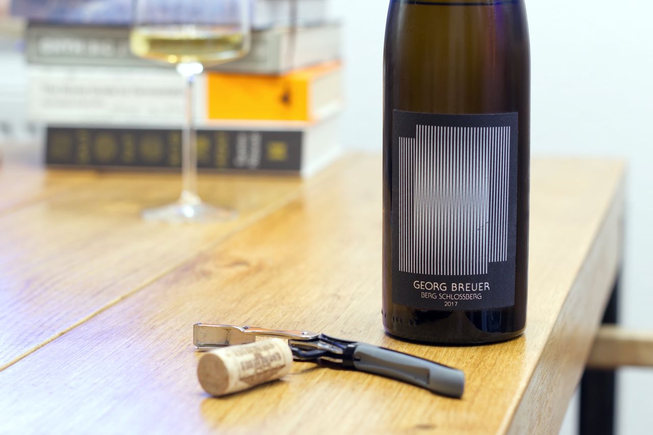 Auf einem Holztisch steht eine Flasche Berg Schlossberg 2017 von Georg Breuer mit Künstleretikett. Im Hintergrund sind ein Weinglas und ein Bücherstapel zu sehen, im Vordergrund liegt der Korken am Kellnermesser.