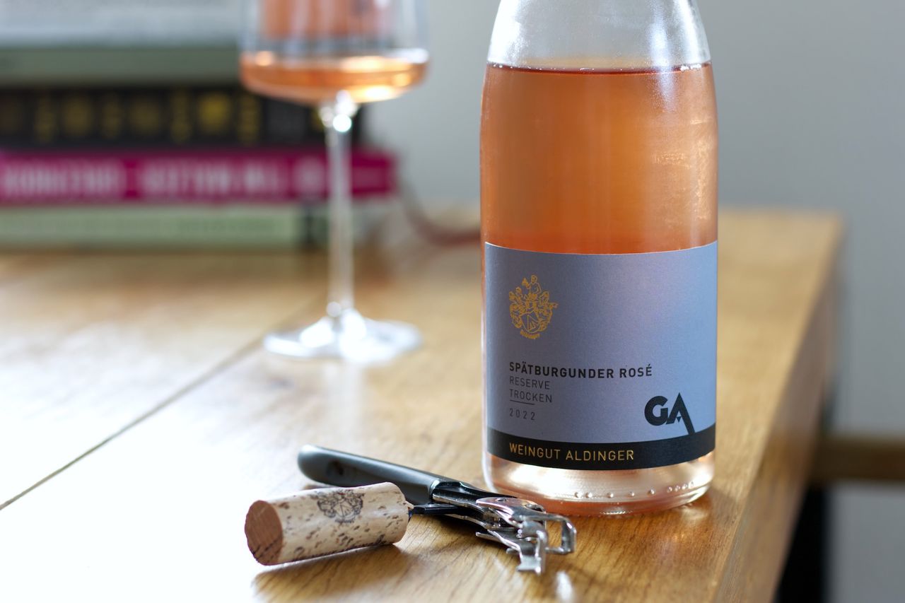 Auf einem Holztisch steht eine Flasche Spätburgunder Rosé Reserve von Aldinger. Im Hintergrund sind ein Weinglas und ein Bücherstapel zu sehen, im Vordergrund liegt der Korken am Kellnermesser.