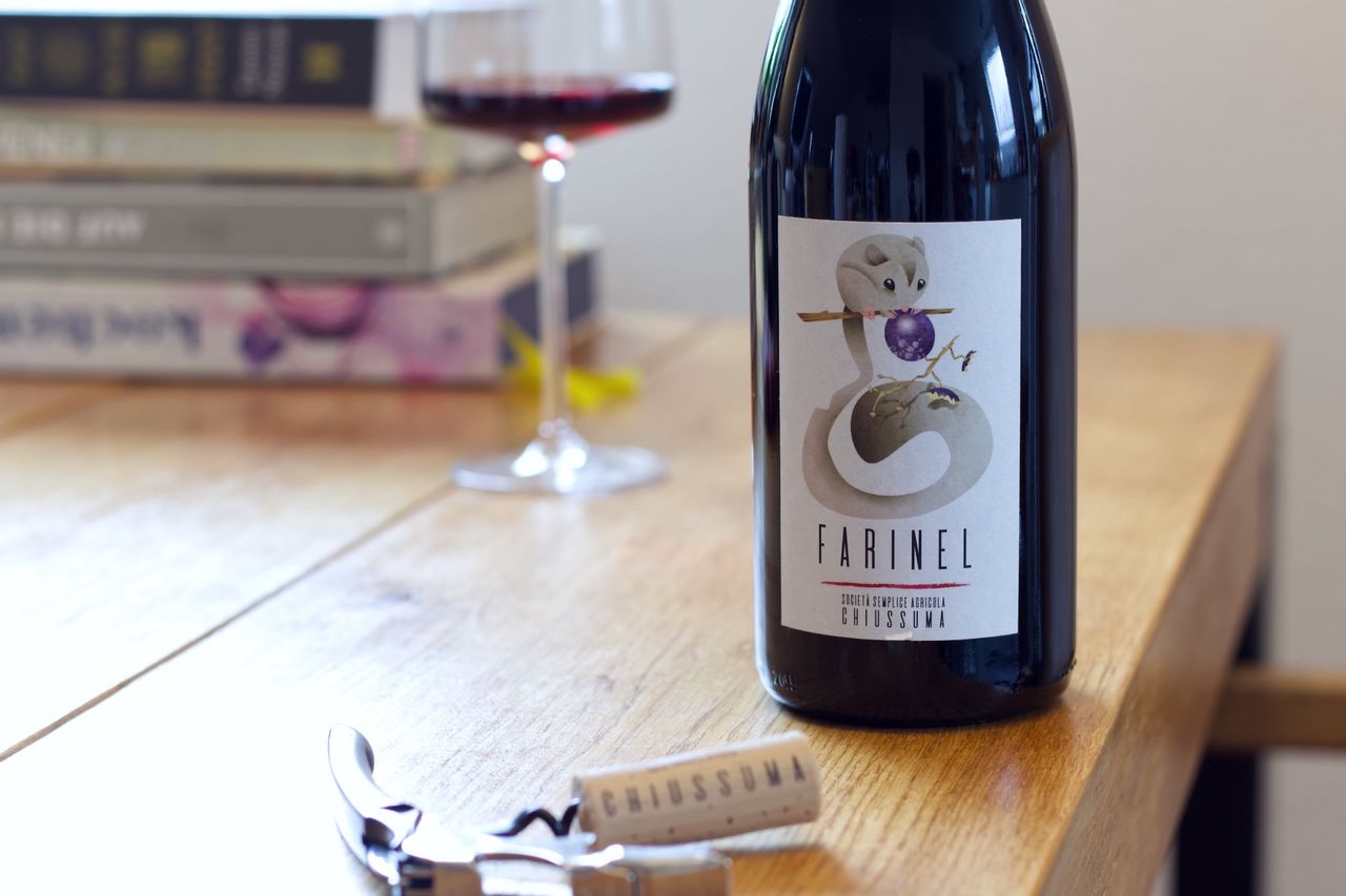 Auf einem Holztisch steht eine Flasche Farinel vom Weingut Chiussuma. Im Hintergrund sind ein Weinglas und ein Bücherstapel zu sehen, im Vordergrund liegt der Korken am Kellnermesser.