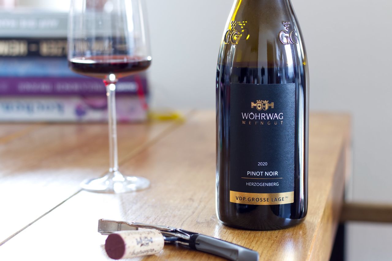 Auf einem Holztisch steht eine Flasche Herzogenberg Pinot Noir vom Weingut Wöhrwag. Im Hintergrund sind ein Weinglas und ein Bücherstapel zu sehen, im Vordergrund liegt der Korken am Kellnermesser.