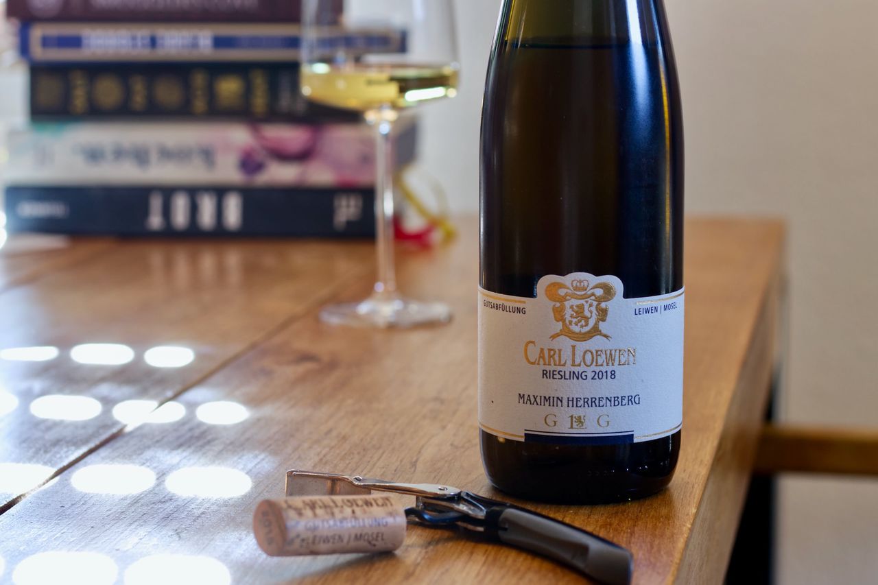 Auf einem Holztisch steht eine Flasche Riesling Maximin Herrenberg vom Weingut Carl Loewen von der Mosel. Im Hintergrund sind ein Weinglas und ein Bücherstapel zu sehen, im Vordergrund liegt der Korken am Kellnermesser.