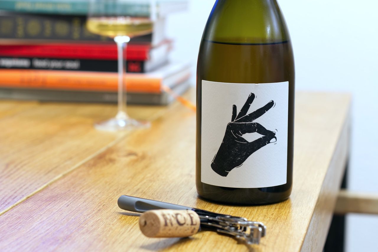Auf einem Holztisch steht eine Flasche Pattaya vom Weingut Vin Noé aus dem Burgund. Auf dem schwarz-weiß gehaltenen Etikett reibt eine Hand zwei Finger aneinander. Im Hintergrund sind ein Weinglas und ein Bücherstapel zu sehen, im Vordergrund liegt der Korken am Kellnermesser.