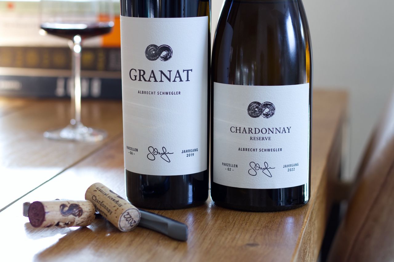 Auf einem Holztisch stehen zwei Flaschen Wein vom Weingut Albrecht Schwegler. Im Hintergrund sind ein Weinglas und ein Bücherstapel zu sehen, im Vordergrund liegen die beiden Korken und ein Kellnermesser.