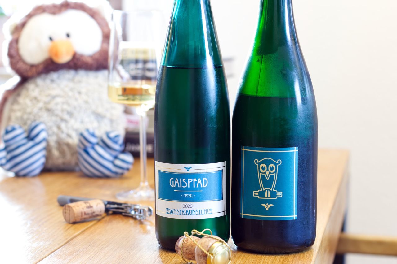 Auf einem Holztisch stehen zwei Flaschen Wein von Weiser-Künstler. Ein Gaispfad Riesling aus 2020 und ein Riesling Sekt mit einer Eule auf dem Etikett. Im Hintergrund sind ein Weinglas und ein Bücherstapel und ein Eulenstofftier zu sehen, im Vordergrund liegt ein Korken am Kellnermesser.