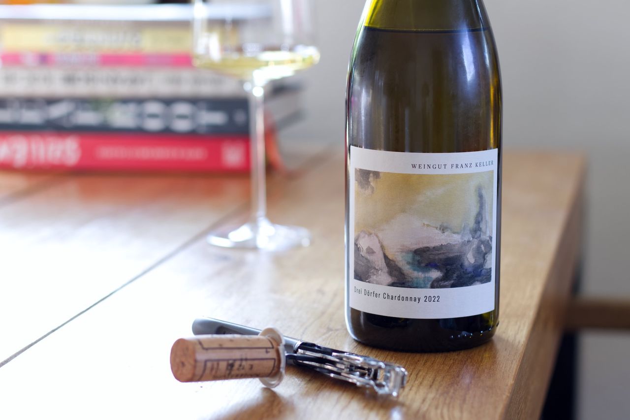 Auf einem Holztisch steht eine Flasche Drei Dörfer von Franz Keller. Im Hintergrund sind ein Weinglas und ein Bücherstapel zu sehen, im Vordergrund liegt der Korken am Kellnermesser.