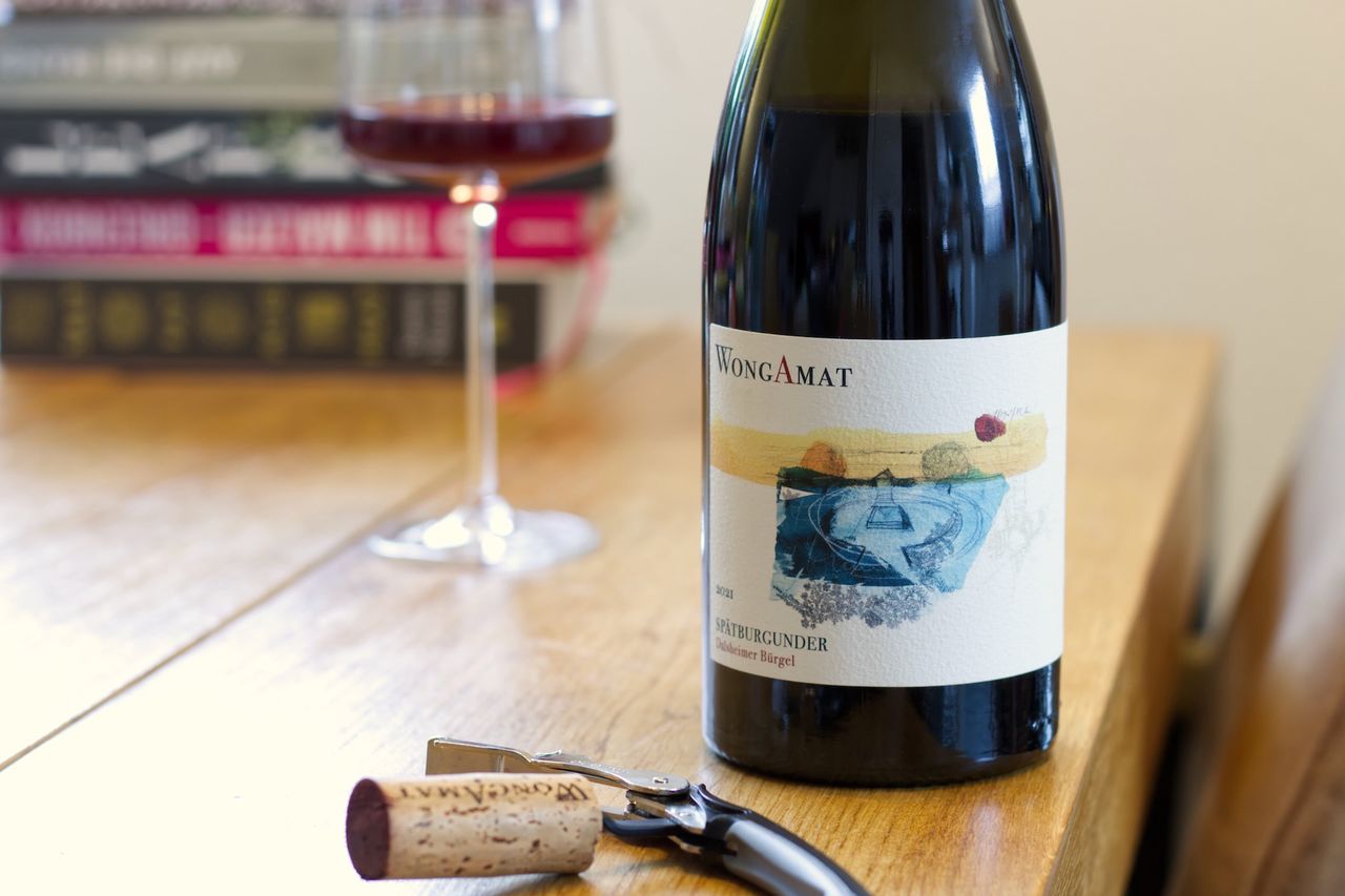 Auf einem Holztisch steht eine Flasche Spätburgunder von WongAmat aus dem Dalsheimer Bürgel. Im Hintergrund sind ein Weinglas und ein Bücherstapel zu sehen, im Vordergrund liegt der Korken am Kellnermesser.