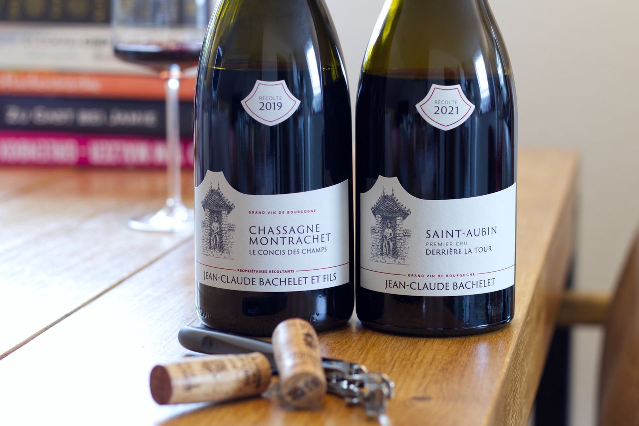 Auf einem Holztisch stehen zwei Flaschen Rotwein von Jean-Claude Bachelet. Im Hintergrund sind ein Weinglas und ein Bücherstapel zu sehen, im Vordergrund liegen die beiden Korken und ein Kellnermesser.