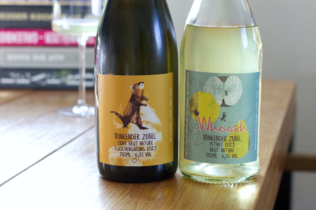 Auf einem Holztisch stehen zwei Flaschen Trinkender Zobel Fruchtschaumwein. Eines der Etiketten zeigt den namensgebenden Zobel. Im Hintergrund sind ein Weinglas und ein Bücherstapel zu sehen.