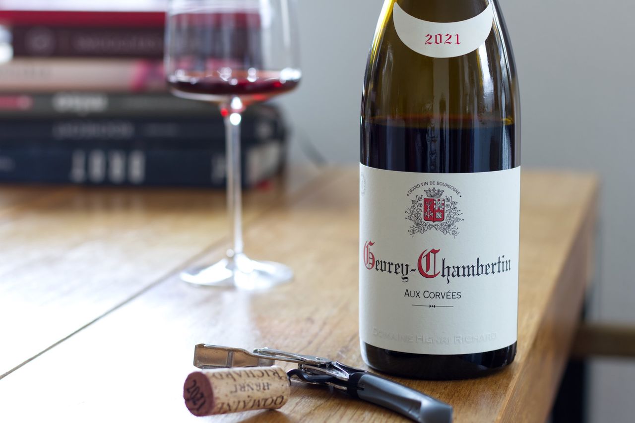 Auf einem Holztisch steht eine Flasche Aux Corvées 2021 vom Weingut Henri Richard. Im Hintergrund sind ein Weinglas und ein Bücherstapel zu sehen, im Vordergrund der Korken am Kellnermesser.