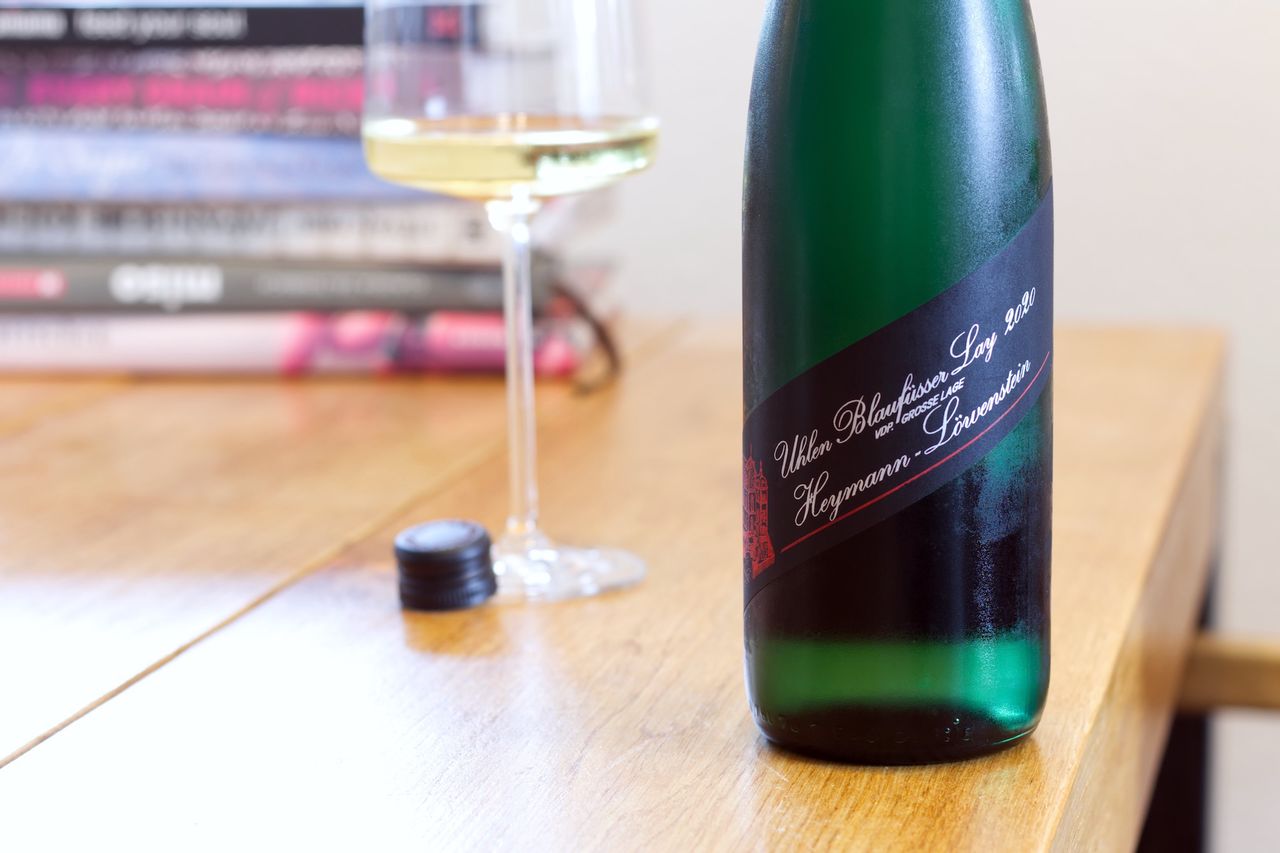 Auf einem Holztisch steht eine Flasche Uhlen Blaufüsser Lay 2020 Riesling von Heymann-Löwenstein. Im Hintergrund sind ein Weinglas und ein Bücherstapel zu sehen.