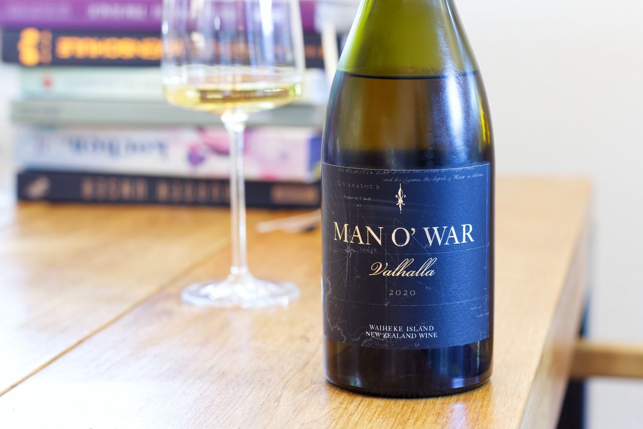 Auf einem Holztisch steht eine Flasche Valhalla Chardonnay 2020 vom Weingut Man O' War. Im Hintergrund sind ein Weinglas und ein Bücherstapel zu sehen.