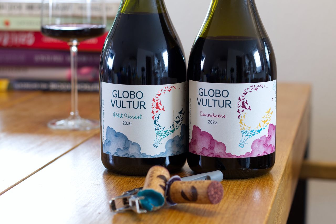 Auf einem Holztisch stehen zwei Flaschen Rotwein von Globo Vultur, ein Petit Verdot und ein Carménère, beide mit einem Heißluftballon auf dem Etikett. Im Hintergrund sind ein Weinglas und ein Bücherstapel zu sehen, im Vordergrund liegen die beiden Korken und ein Kellnermesser.
