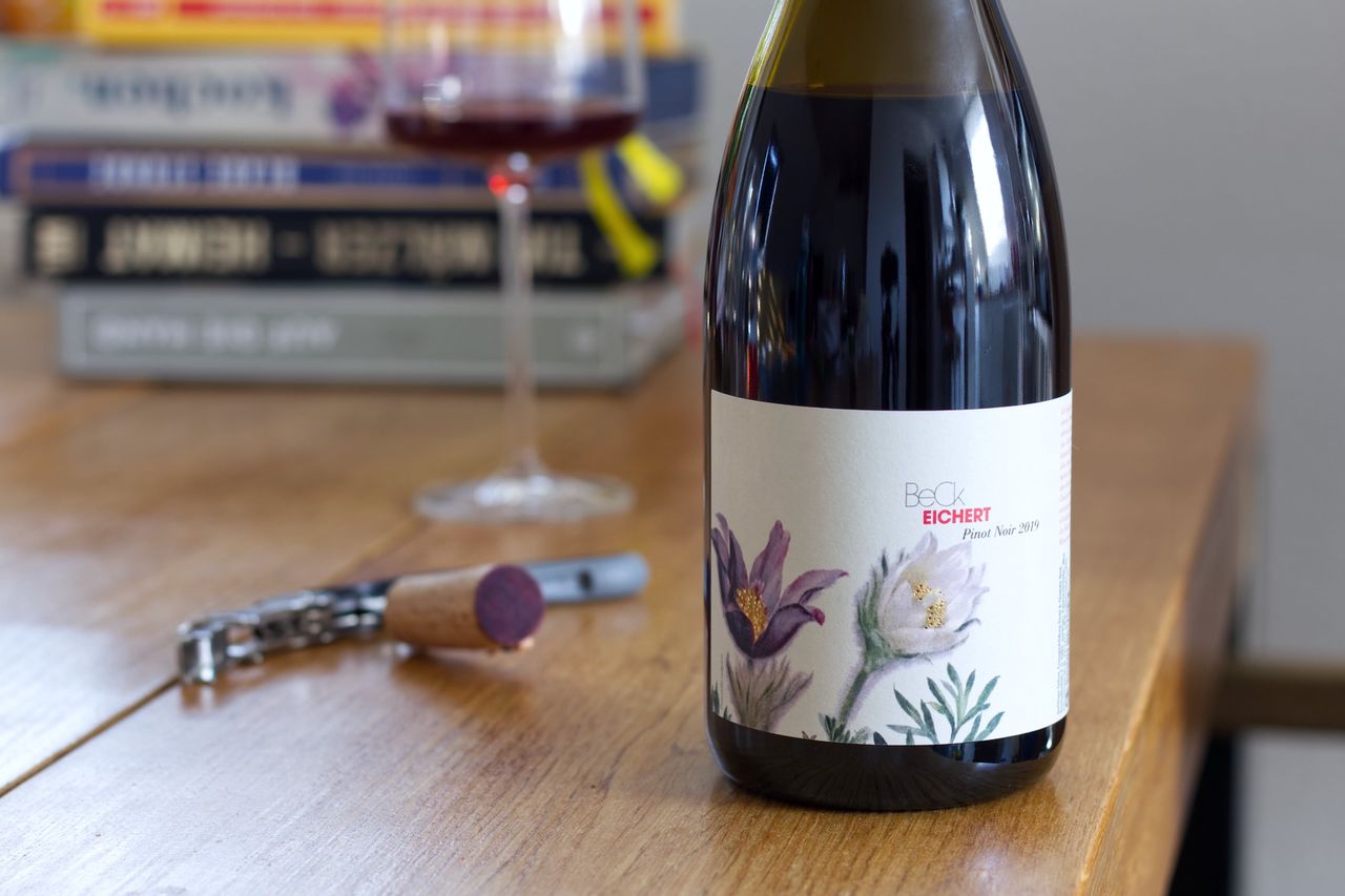 Auf einem Holztisch steht eine Flasche Spätburgunder von Beck mit Blumen auf dem Etikett. Im Hintergrund sind ein Weinglas und ein Bücherstapel zu sehen, im Vordergrund liegt der Korken am Kellnermesser.