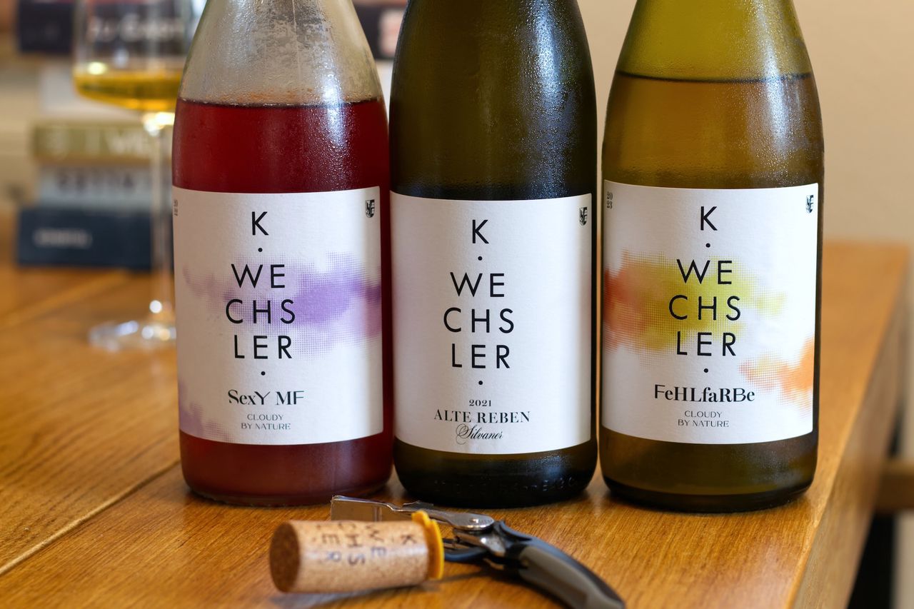Drei Flaschen Wein vom Weingut Wechsler stehen auf einem Holztisch. Die Etiketten sind schlicht mit schwarzer Schrift und ein paar Farbklecksen. Davor liegt ein Korken am Kellnermesser, dahinter steht ein Weinglas und ein Stapel Bücher.