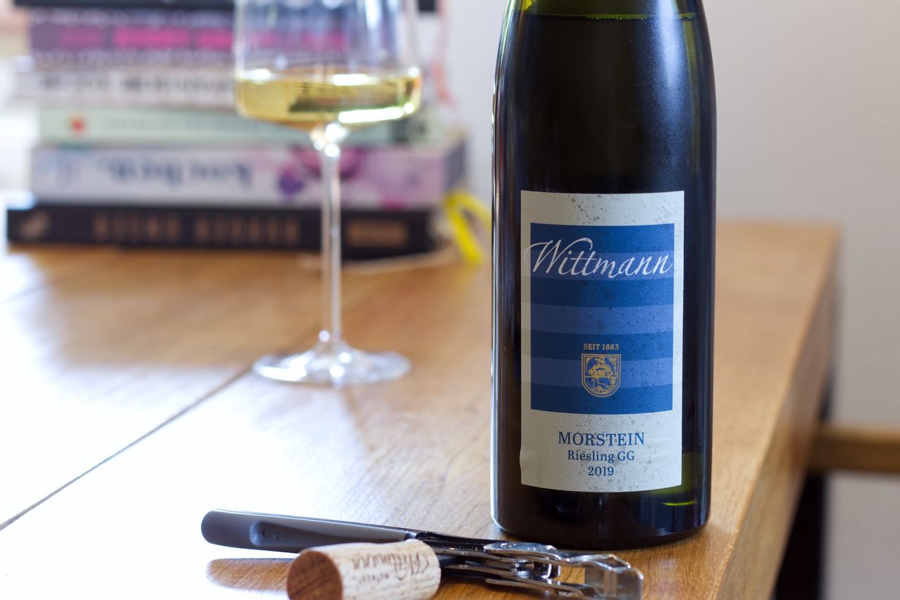 Auf einem Holztisch steht eine Flasche Riesling von Wittmann mit dem blau-weißen Etikett. Im Hintergrund sind ein Weinglas und ein Bücherstapel zu sehen, im Vordergrund liegt der Korken am Kellnermesser.