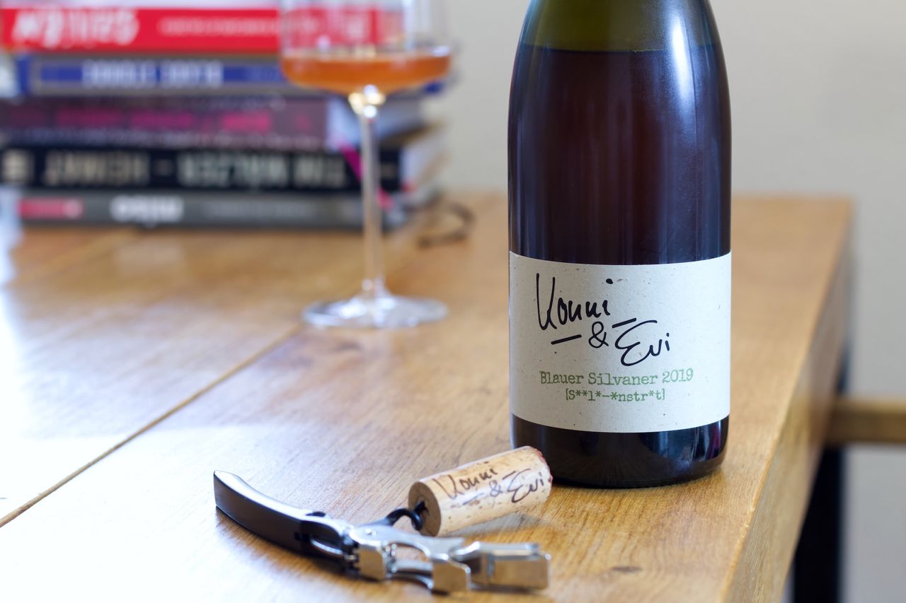 Eine Flasche Silvaner von Konni und Evi steht auf einem Holztisch. Im Vordergrund liegt der Korken der Flasche am Kellnermesser. Hinter der Flasche stehen ein Weinglas und ein Stapel Bücher.