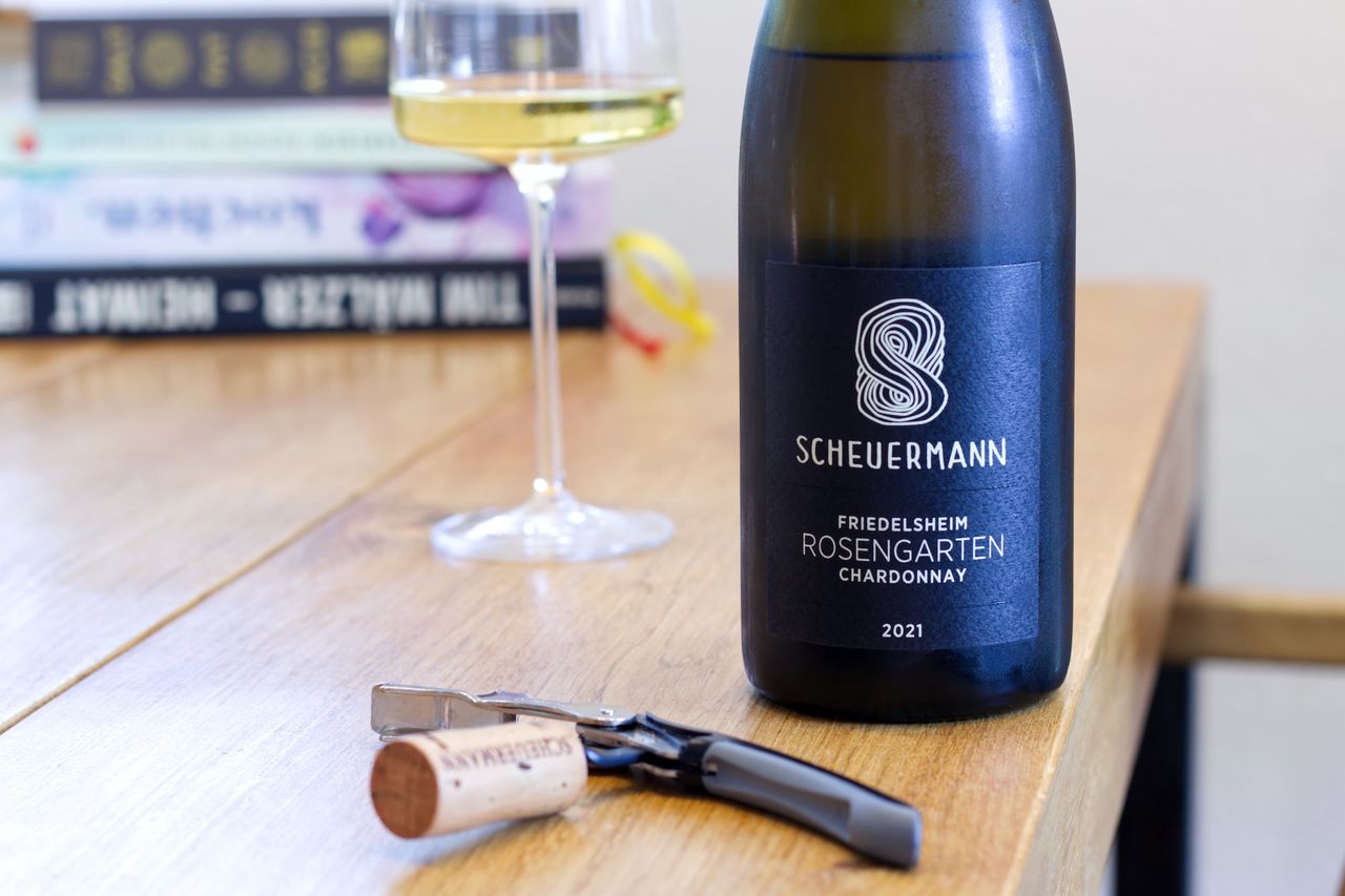 Eine Flasche Chardonnay aus dem Rosengarten vom Weingut Scheuermann steht auf einem Holztisch. Im Vordergrund liegt der Korken der Flasche am Kellnermesser. Hinter der Flasche stehen ein Weinglas und ein Stapel Bücher.