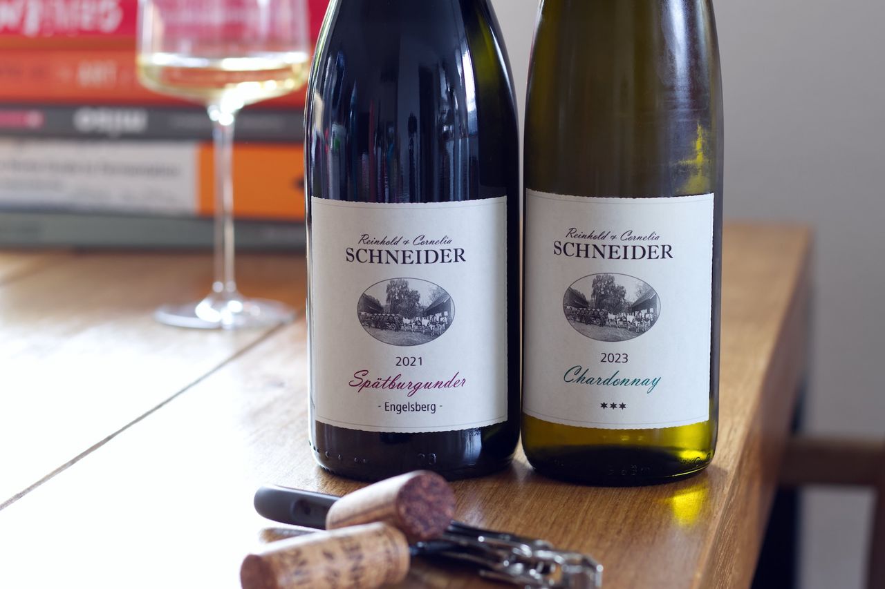 Zwei Flaschen Wein vom Weingut Reinhold und Cornelia Schneider stehen auf einem Holztisch. Im Vordergrund liegen die beiden Korken und ein Kellnermesser. Dahinter stehen ein Weinglas und ein Stapel Bücher.