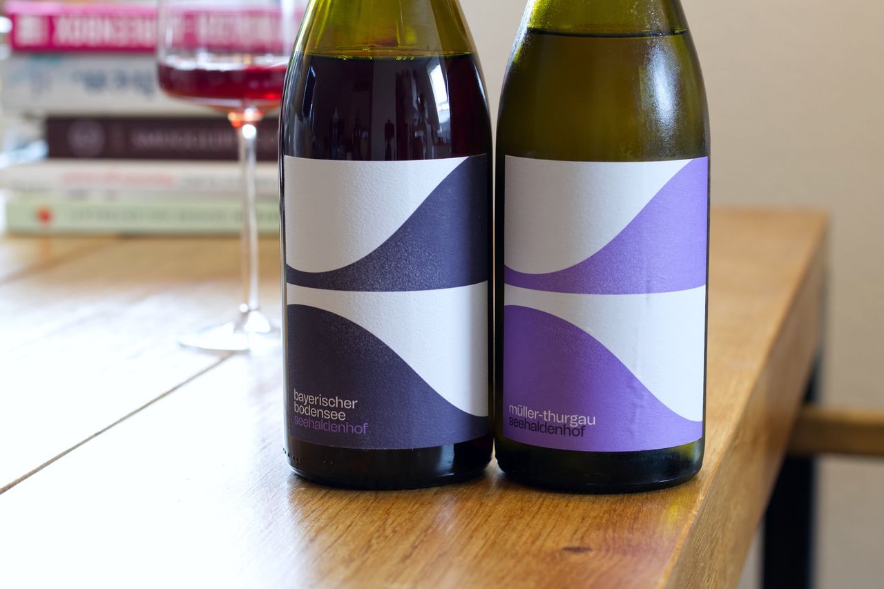 Zwei Flaschen Wein vom Weingut Seehaldenhof stehen auf einem Holztisch. Dahinter stehen ein Weinglas und ein Stapel Bücher.