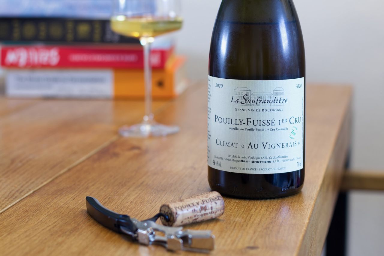 Eine Flasche Chardonnay Au Vignerais mit einem kleinen grünen Zen Stempel von La Soufrandière steht auf einem Holztisch. Im Vordergrund liegt der Korken am Kellnermesser. Dahinter stehen ein Weinglas und ein Stapel Bücher.