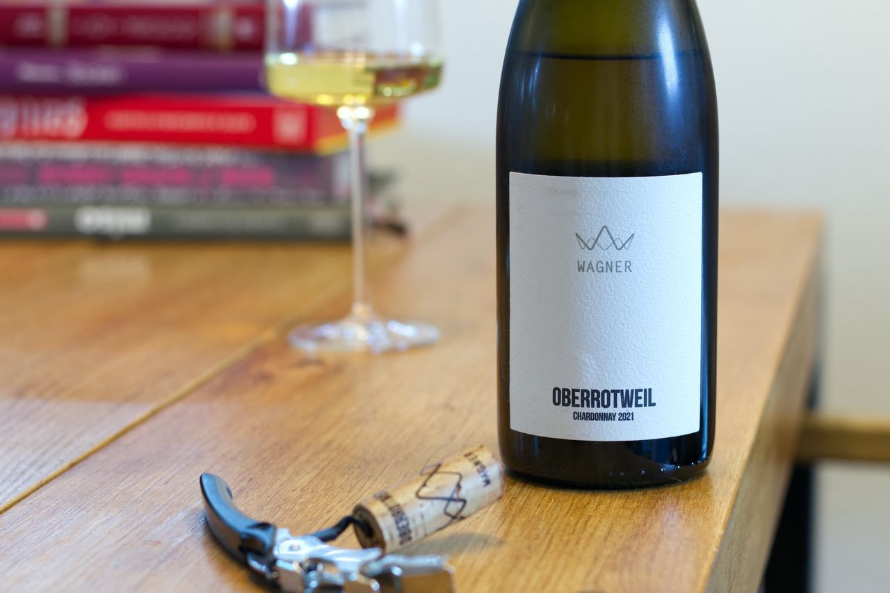 Auf einem Holztisch steht eine Flasche Chardonnay vom Weingut Peter Wagner. Im Hintergrund sind ein Weinglas und ein Bücherstapel zu sehen. Vor der Flasche liegt der Korken am Kellnermesser.