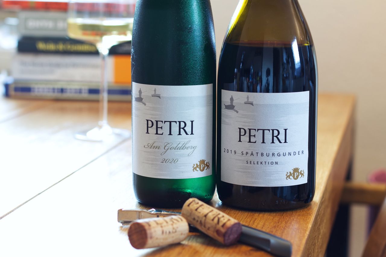 Zwei Flaschen Wein vom Weingut Petri stehen auf einem Holztisch. Auf den Etiketten sind Angler abgebildet. Dahinter stehen ein Weinglas und ein Stapel Bücher. Vor den Flaschen liegen die beiden Korken und ein Kellnermesser.