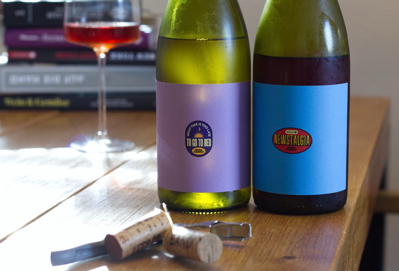 Auf einem Holztisch stehen zwei Flaschen von Wein Goutte. Die Labels sind schlicht und knallig zu gleich in intensiven Farben mit kleinem Logo. Im Hintergrund sind ein Weinglas und ein Bücherstapel zu sehen. Vor der Flasche liegen Korken und Kellnermesser.