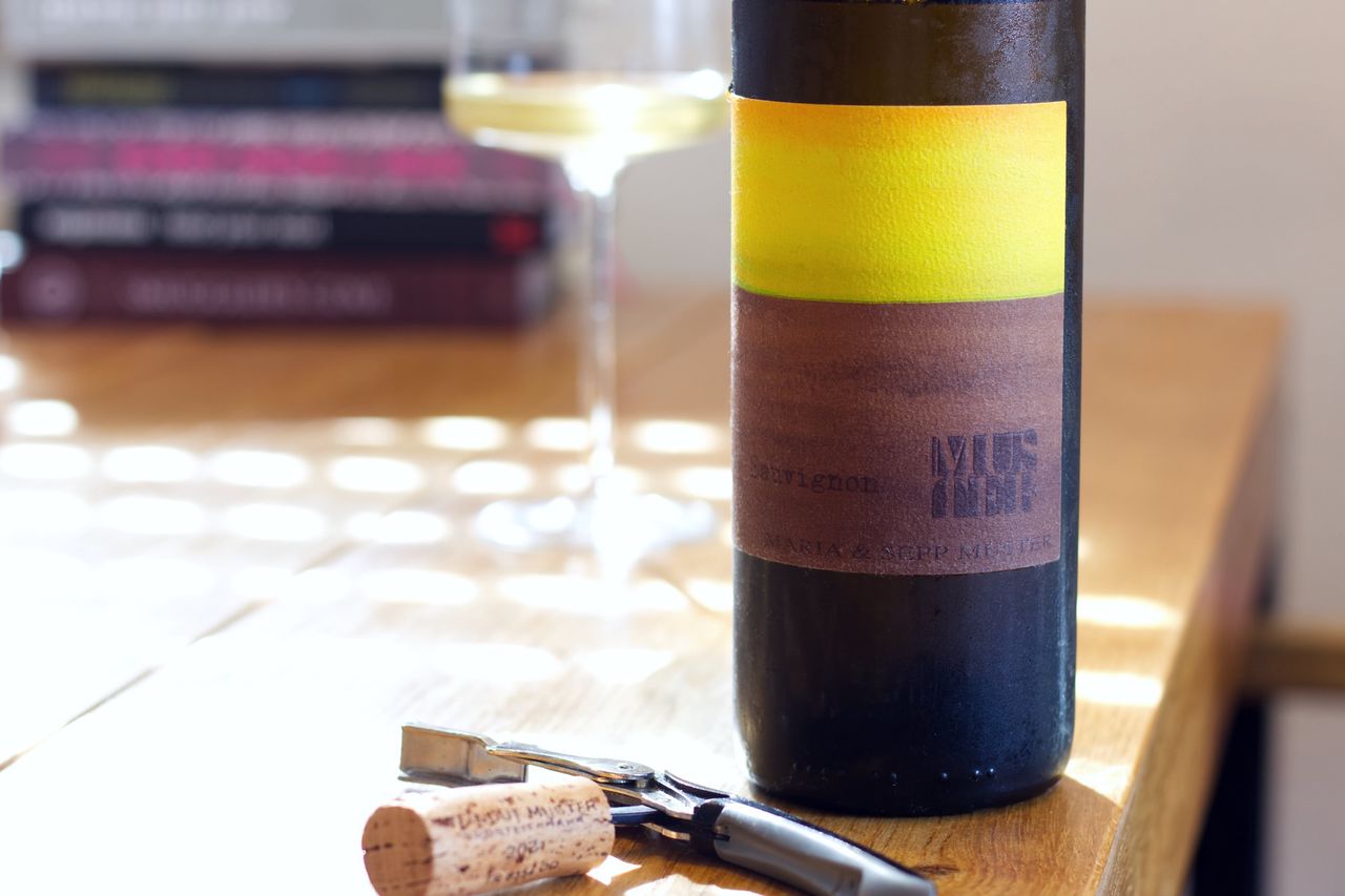 Eine Flasche Sauvignon 2021 von Maria und Sepp Muster steht auf einem Holztisch. Im Vordergrund liegt der Korken der Flasche am Kellnermesser. Hinter der Flasche stehen ein Weinglas und ein Stapel Bücher.