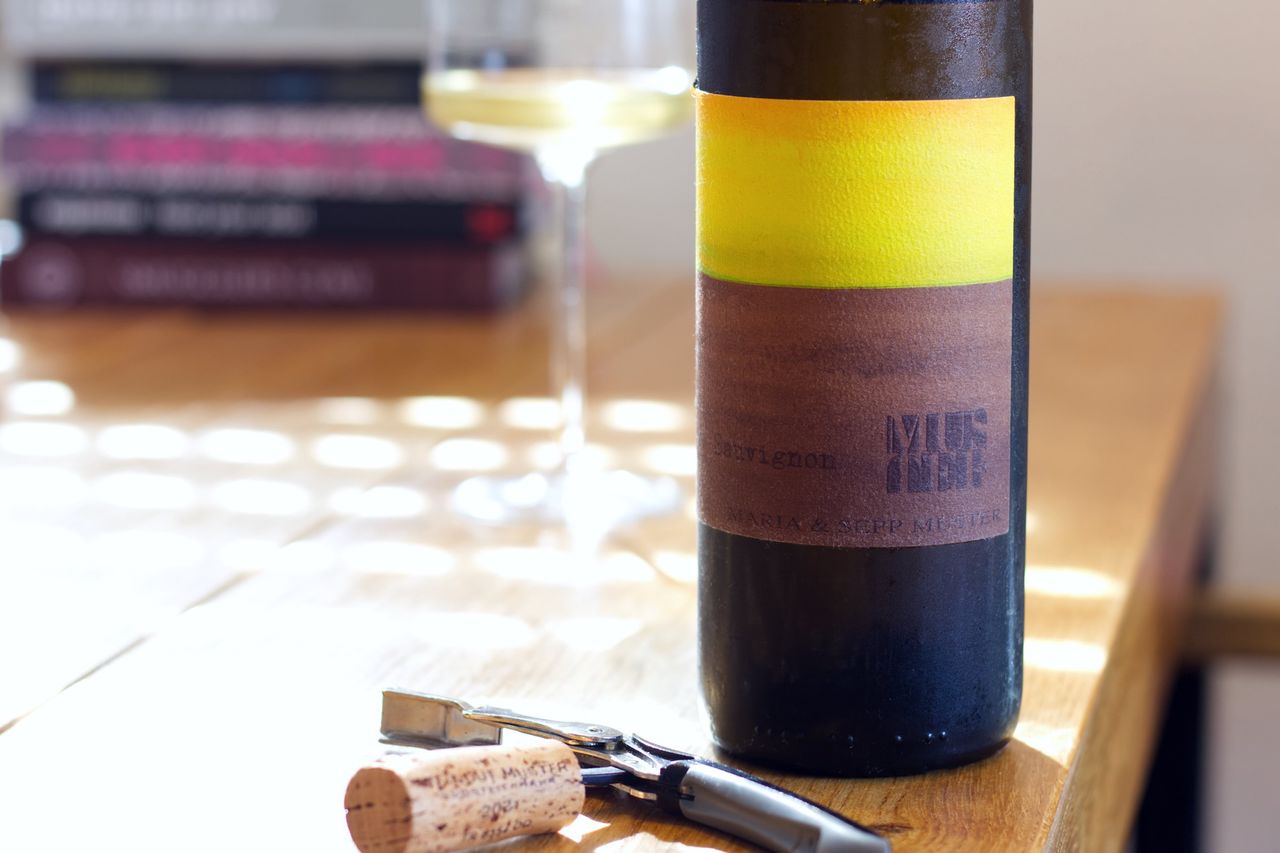 Eine Flasche Sauvignon 2021 von Maria und Sepp Muster steht auf einem Holztisch. Im Vordergrund liegt der Korken der Flasche am Kellnermesser. Hinter der Flasche stehen ein Weinglas und ein Stapel Bücher.