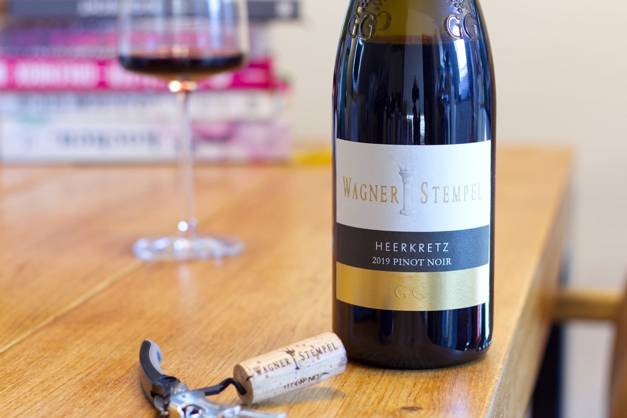 Auf einem Holztisch steht eine Flasche Pinot Noir aus der Heerkretz von Wagner-Stempel. Im Hintergrund sind ein Weinglas und ein Bücherstapel zu sehen. Vor der Flasche liegt der Korken am Kellnermesser.