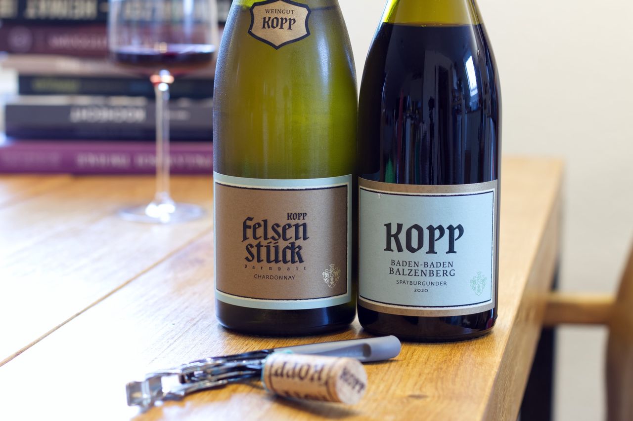Auf einem Holztisch stehen zwei Flaschen Wein vom Weingut Kopp. Im Hintergrund sind ein Weinglas und ein Bücherstapel zu sehen. Vor der Flasche liegen Korken und Kellnermesser.
