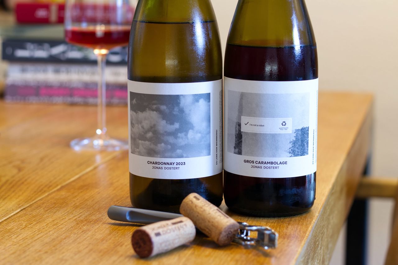 Auf einem Holztisch stehen zwei Flaschen Wein von Jonas Dostert. Im Hintergrund sind ein Weinglas und ein Bücherstapel zu sehen. Vor den Flaschen liegen Korken und Kellnermesser.