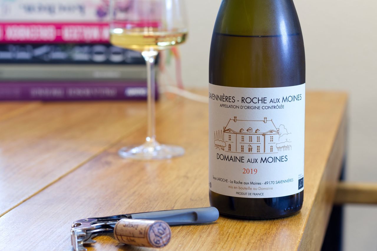 Eine Flasche Roche Aux Moines Chenin Blanc 2019 von der Domaine Aux Moines steht auf einem Holztisch. Dahinter stehen ein Weinglas und ein Stapel Bücher. Vor der Flasche liegt der Korken und ein Kellnermesser.