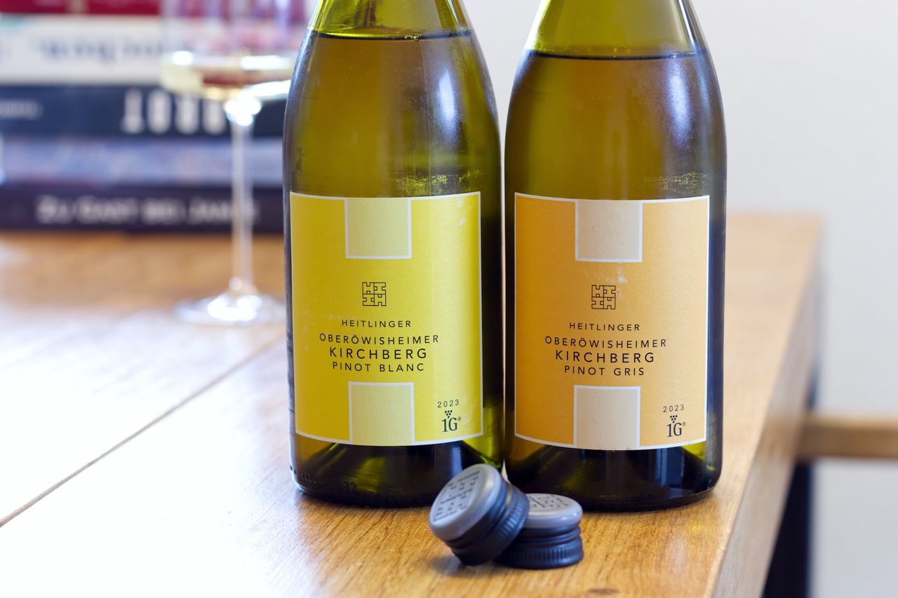 Auf einem Holztisch stehen zwei Flaschen Wein vom Weingut Heitlinger. Im Hintergrund sind ein Weinglas und ein Bücherstapel zu sehen. Vor den Flaschen liegen die Schraubdeckel.