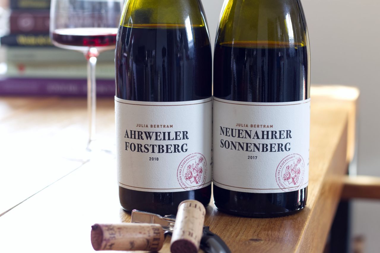 Auf einem Holztisch stehen zwei Flaschen Wein von Julia Bertram. Im Hintergrund sind ein Weinglas und ein Bücherstapel zu sehen. Vor der Flasche liegen Korken und Kellnermesser.