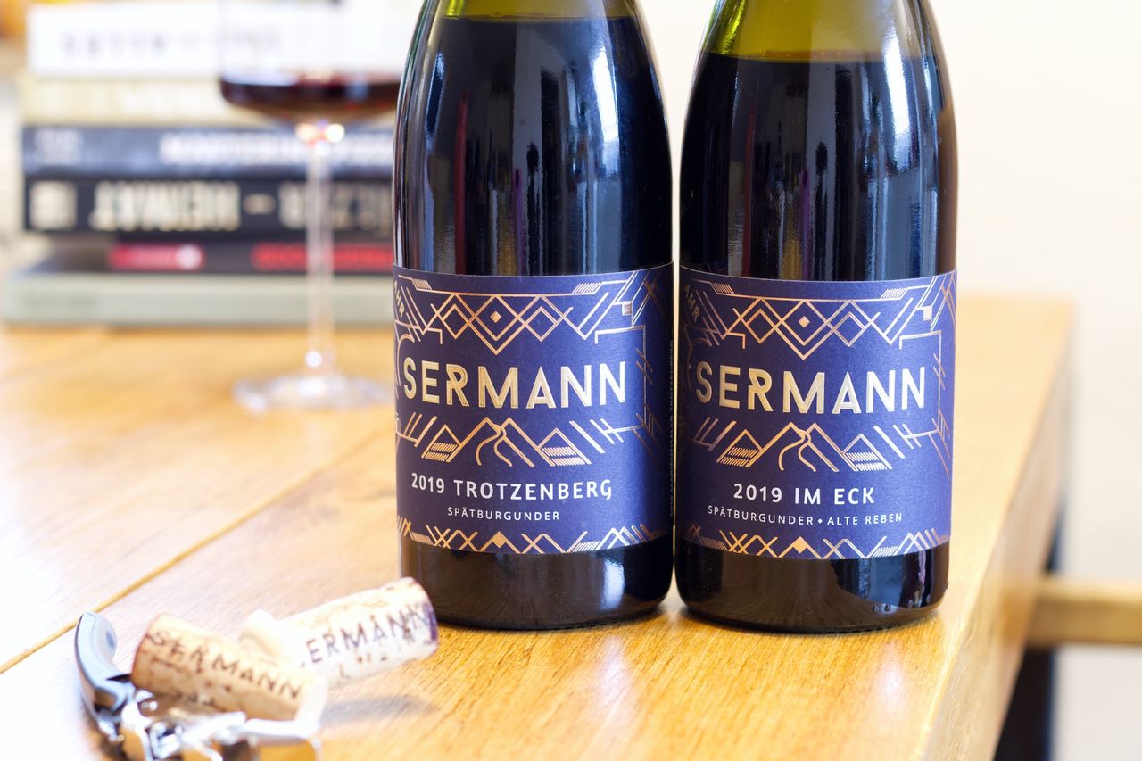 Zwei Flaschen Wein vom Weingut Sermann mit blauem Etikett stehen auf einem Holztisch. Dahinter stehen ein Weinglas und ein Stapel Bücher. Vor den Flaschen liegen die beiden Korken und ein Kellnermesser.
