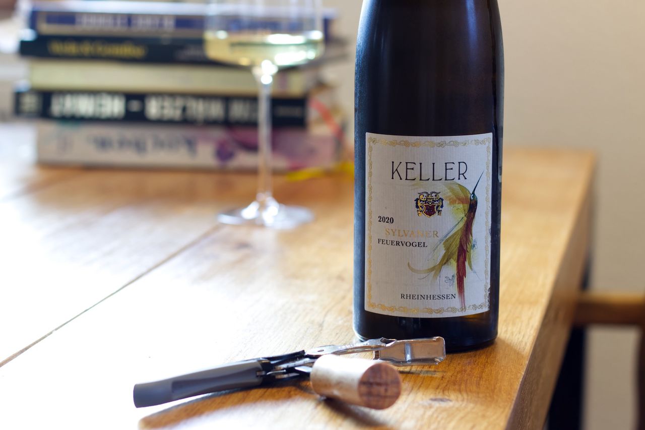 Eine Flasche Feuervogel 2020 vom Weingut Keller steht auf einem Holztisch. Dahinter stehen ein Weinglas und ein Stapel Bücher. Vor den Flaschen liegen der Korken und ein Kellnermesser.