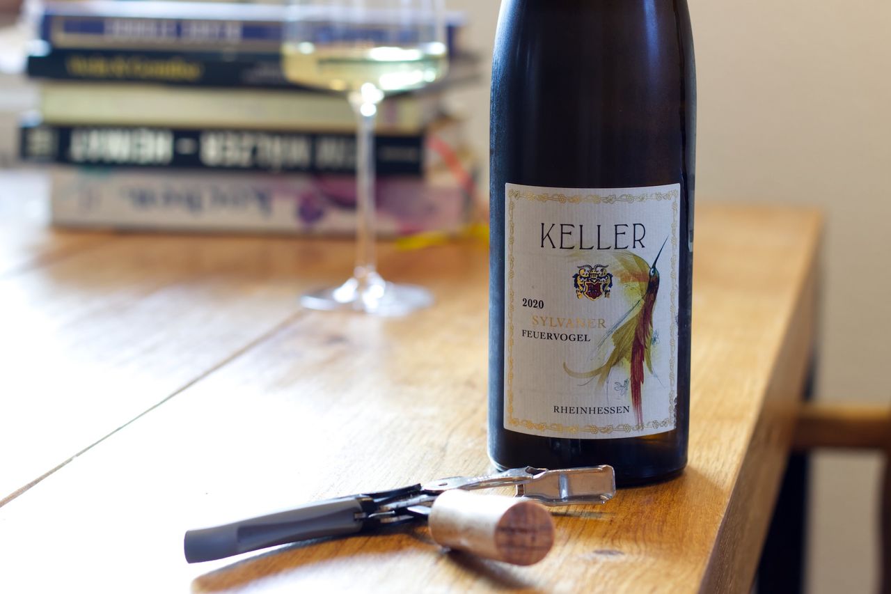Eine Flasche Feuervogel 2020 vom Weingut Keller steht auf einem Holztisch. Dahinter stehen ein Weinglas und ein Stapel Bücher. Vor den Flaschen liegen der Korken und ein Kellnermesser.