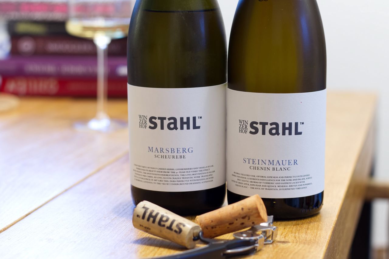 Auf einem Holztisch stehen zwei Flaschen Wein vom Winzerhof Stahl aus Franken. Im Hintergrund sind ein Weinglas und ein Bücherstapel zu sehen. Vor den Flaschen liegen Korken und Kellnermesser.