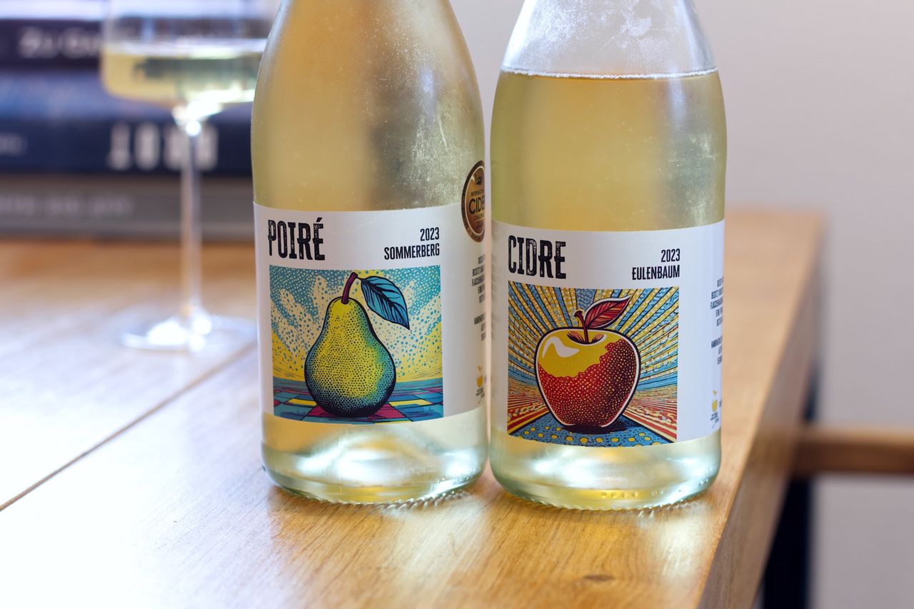 Auf einem Holztisch stehen zwei Flaschen Obstschaumwein von Der Kleine Cidre.Betrieb. Im Hintergrund sind ein Weinglas und ein Bücherstapel zu sehen.