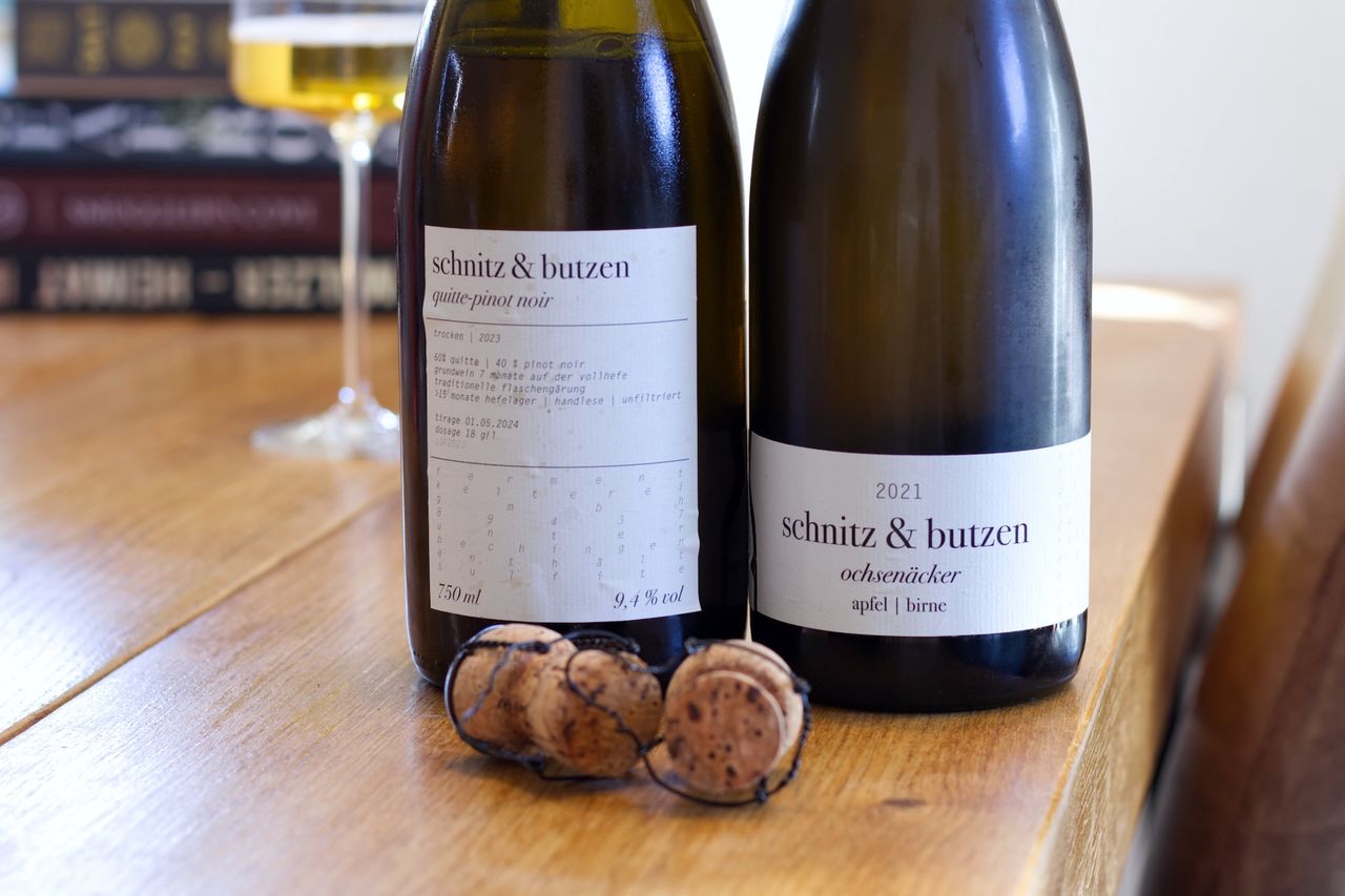 Auf einem Holztisch stehen zwei Flaschen Obstschaumwein von Schnitz und Butzen. Im Hintergrund sind ein Weinglas und ein Bücherstapel zu sehen.