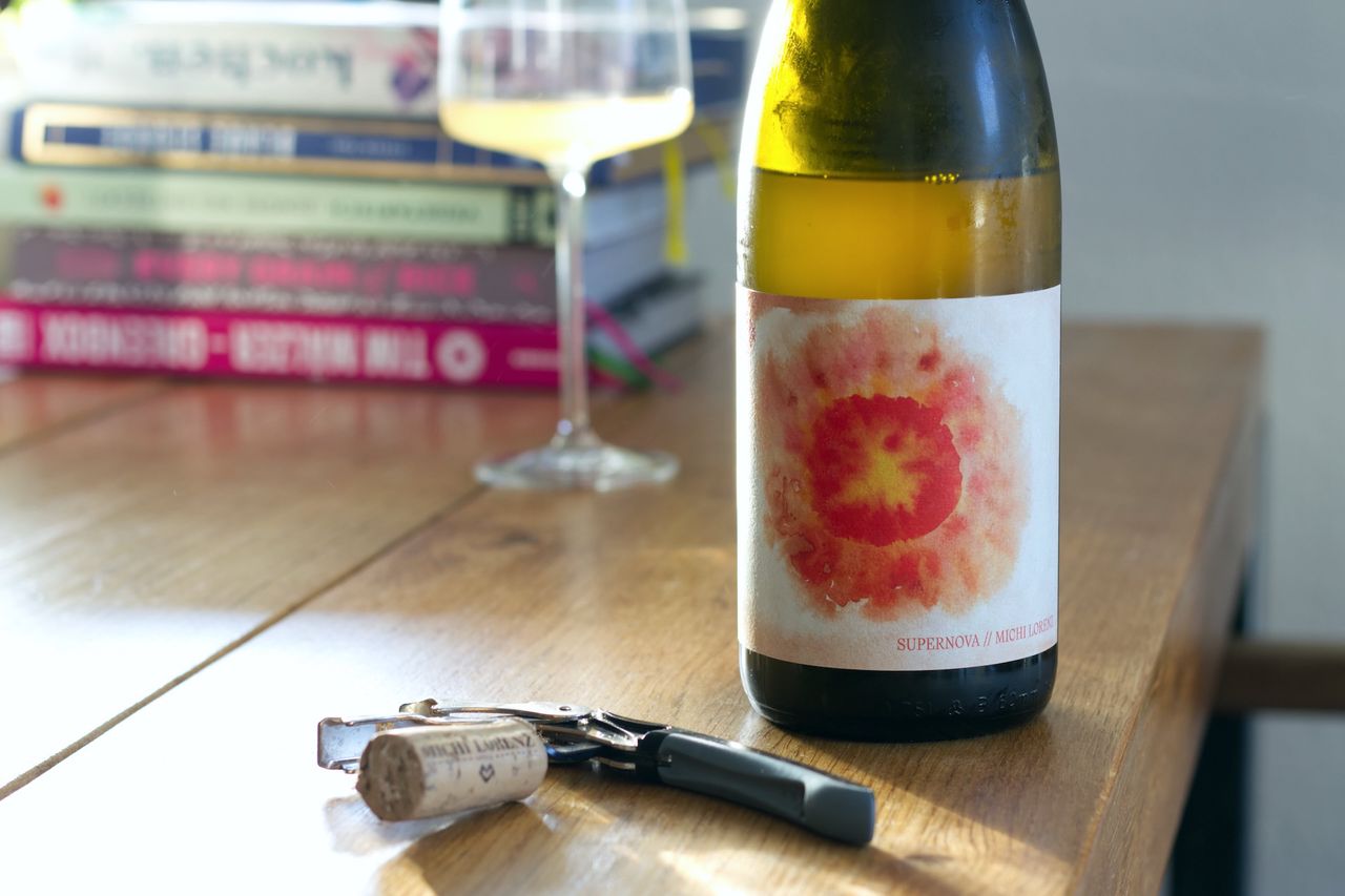 Auf einem Holztisch steht eine Flasche Supernova von Michi Lorenz. Im Hintergrund sind ein Weinglas und ein Bücherstapel zu sehen, vor der Flasche liegt der Korken am Kellnermesser.