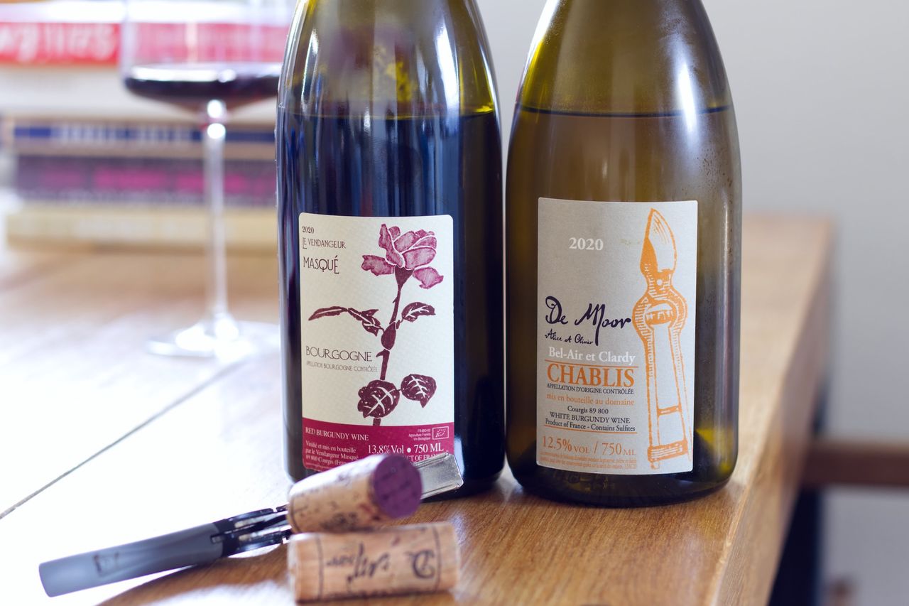 Auf einem Holztisch stehen zwei Flaschen Wein vom Weingut De Moor. Im Hintergrund sind ein Weinglas und ein Bücherstapel zu sehen. Vor den Flaschen liegen die Korken am Kellnermesser.