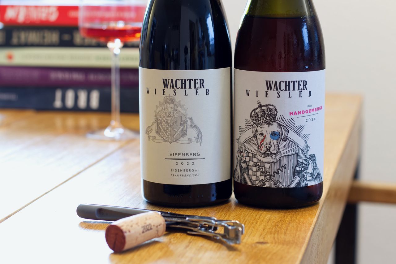 Auf einem Holztisch stehen zwei Flaschen Wein vom Weingut Wachter-Wiesler mit dem Hund auf dem Label. Im Hintergrund sind ein Weinglas und ein Bücherstapel zu sehen. Vor den Flaschen liegt ein Korken am Kellnermesser.