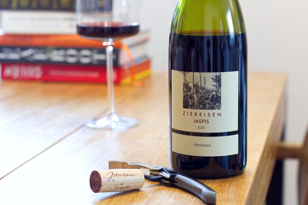 Auf einem Holztisch steht eine Flasche Jaspis Hermann Spätburgunder von Ziereisen. Im Hintergrund sind ein Weinglas und ein Bücherstapel zu sehen, vor der Flasche liegen Korken und Kellnermesser.