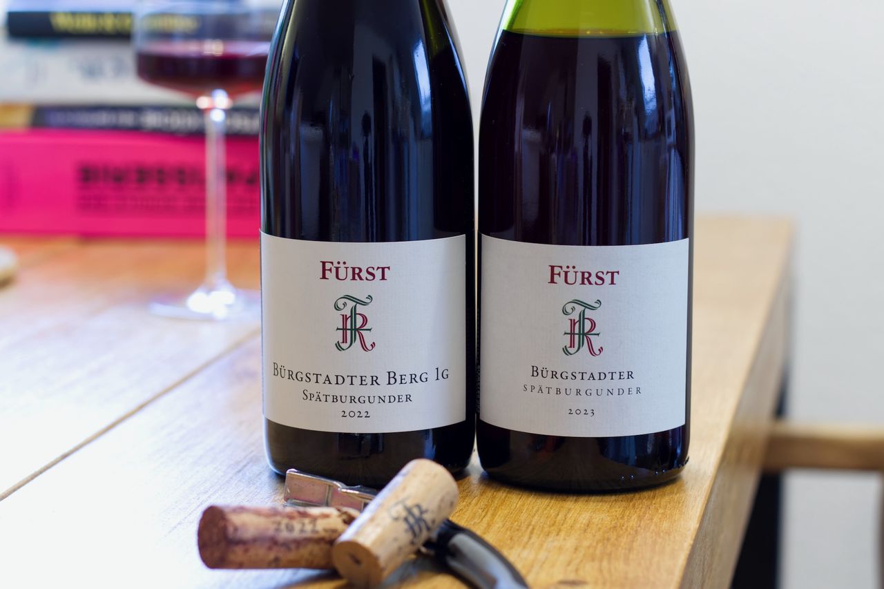Auf einem Holztisch stehen zwei Flaschen Wein vom Weingut Fürst. Im Hintergrund sind ein Weinglas und ein Bücherstapel zu sehen. Vor den Flaschen liegen die Korken am Kellnermesser.