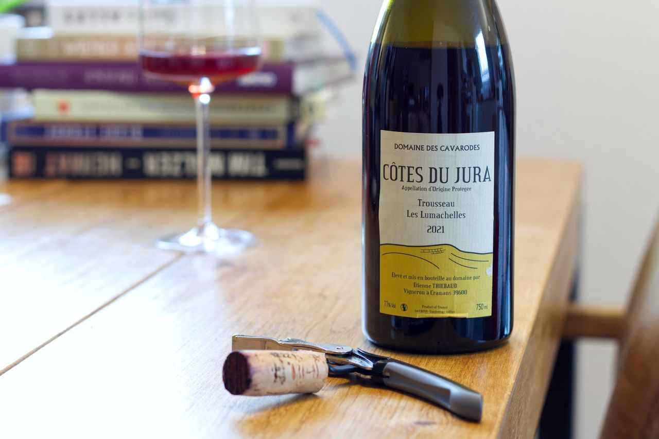 Auf einem Holztisch steht eine Flasche Les Lumachelles von Cavarodes. Im Hintergrund sind ein Weinglas und ein Bücherstapel zu sehen, vor der Flasche liegen Korken und Kellnermesser.