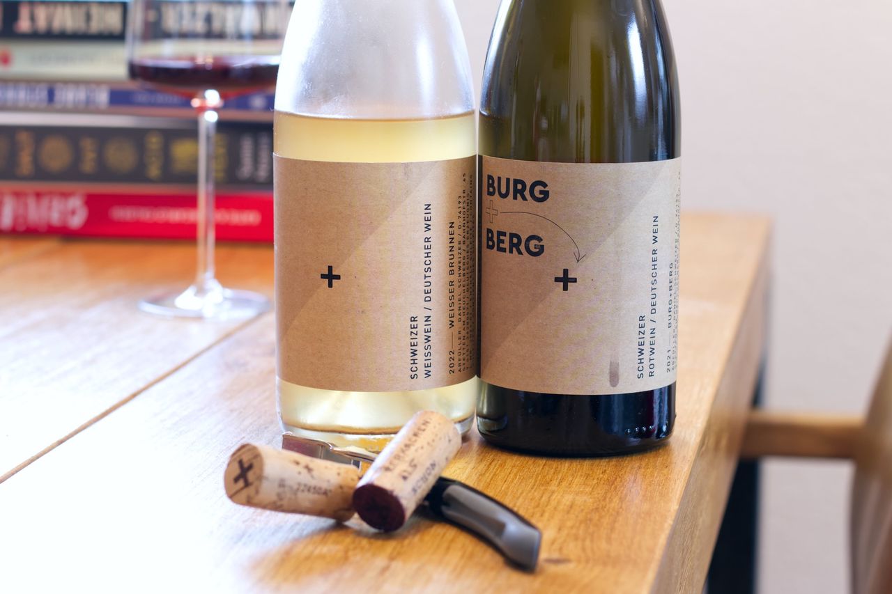 Auf einem Holztisch stehen zwei Flaschen Wein von Daniel Schweizer. Im Hintergrund sind ein Weinglas und ein Bücherstapel zu sehen. Vor den Flaschen liegen die Korken am Kellnermesser.