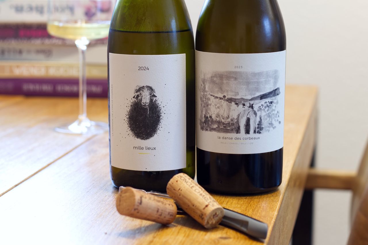 Auf einem Holztisch stehen zwei Flaschen Wein vom Weingut La Grange de l'Oncle Charles. Im Hintergrund sind ein Weinglas und ein Bücherstapel zu sehen. Vor den Flaschen liegt ein Korken am Kellnermesser.